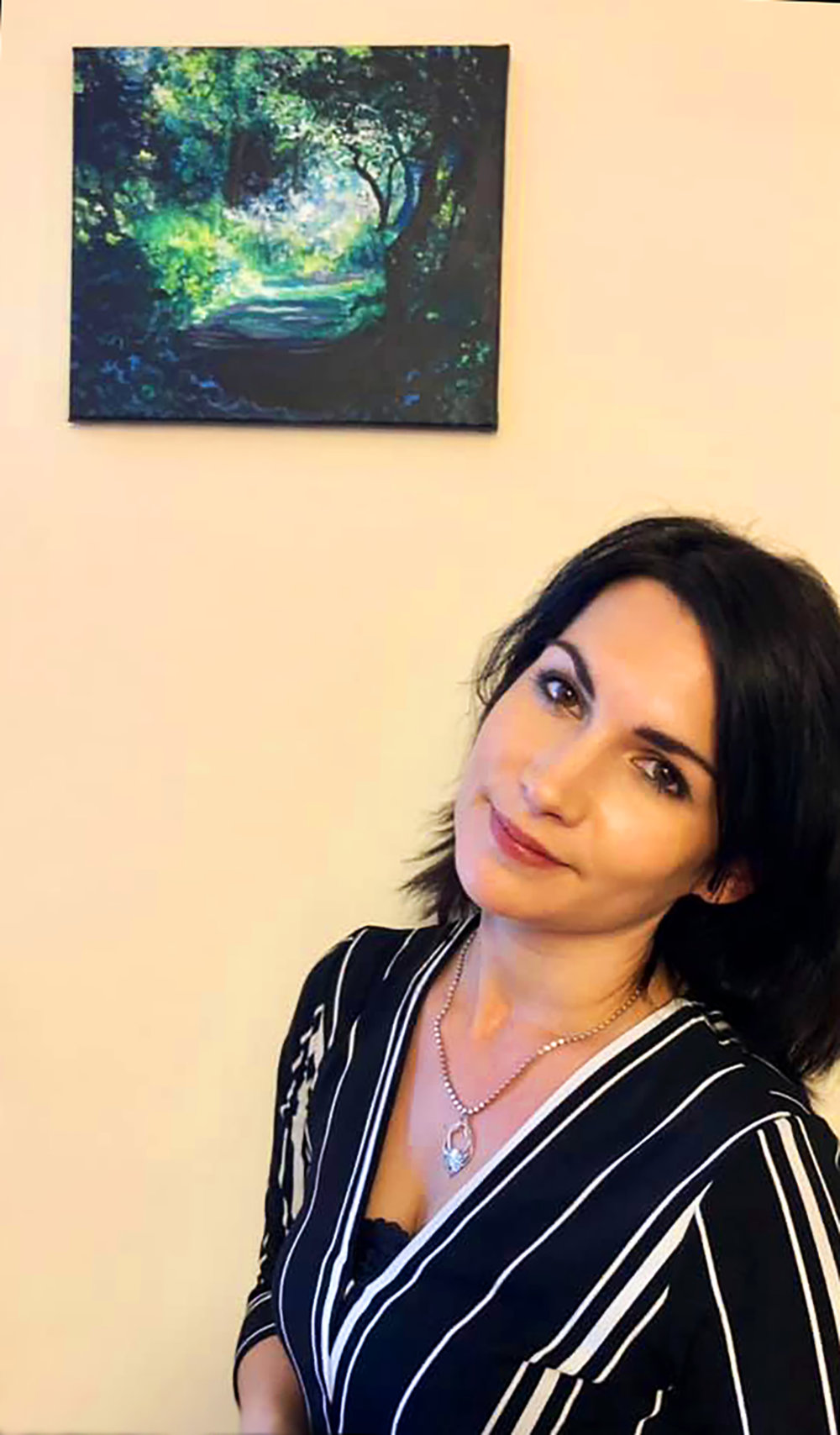 Madeleine Pires beneath her painting of a sunlit woodland path through an arch of trees